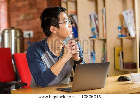 Thoughtful young asian man in glasses using laptop in cafe