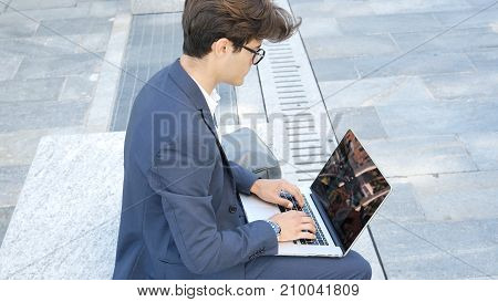 Handsome young man sitting on a stone bench working outdoors in an urban setting typing information onto his laptop computer