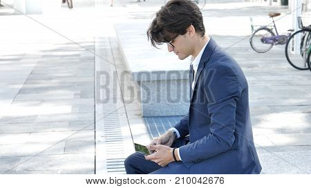 Handsome eleganty young business man wearing jacket sitting and working, looking down at a tablet computer that he is holding, outdoor