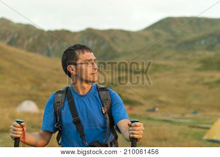 a man with a backpack in a blue T-shirt and glasses with sticks for Nordic walking is photographed by belt against the background of a blurry mountain valley