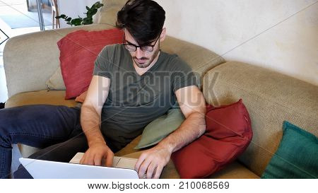 Attractive Young Man with Serious Expression, with Laptop on Couch, Working on his Start-up Business - Young Male College or University Student Doing Homework on Sofa