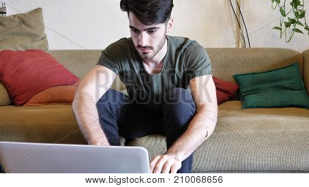 Attractive Young Man with Serious Expression, with Laptop on Couch, Working on his Start-up Business - Young Male College or University Student Doing Homework on Sofa