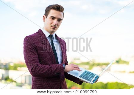 Serious Businessman In Red Suit And Shirt With Tie Stand On The Roof With Laptop