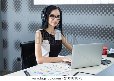 Beautiful Young Businesswoman In Black Dress, Headphones And Glasses Sit At The Table And Work On La