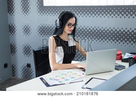 Beautiful Young Businesswoman In Black Dress, Headphones And Glasses Sit At The Table And Work On La