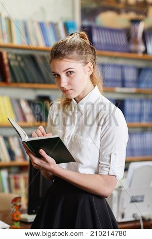 Pretty smiling student reading the book in the library. Beautiful girl posing on the camera and holding book.Shelves with books on the background. Close-up of girl.