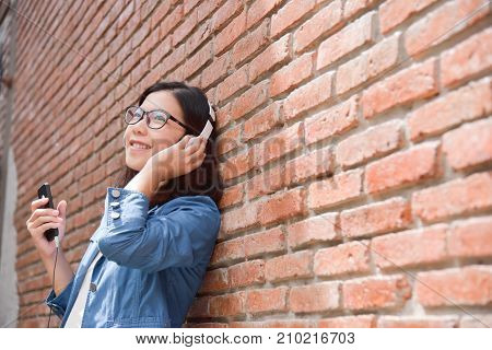 Happy Beautiful Asian woman in blue shirt listening to the music with headphone via her smart phone on old red brick wall background. copy space.