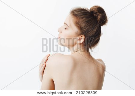Bauty and health. Good-looking thin healthy caucasian girl with dark hair in bun hairstyle and naked skin, turns with back in camera, touching shoulders with hands, having relaxed and sutisfied look with closed eyes