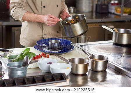 Chef put out with spoon stewing vegetables. Cropped image of male chef going putting garnishing into stainless bowl. Food preparation at professional kitchen.