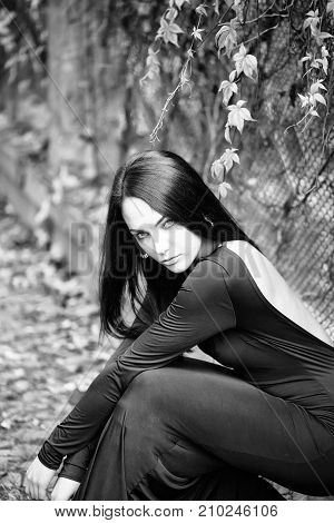 One sexual young attractive brunette woman with long hair sitting in forest near fense with autumn leaves plant in black dress outdoor on natural background vertical picture