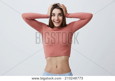 Close up portrait of beautiful joyful young caucaian girl with dark long hair in pink stylish top and blue jeans smiling with teeth, holding head with hands, looking in camera with happy and excited expression after receiving present for Christmas