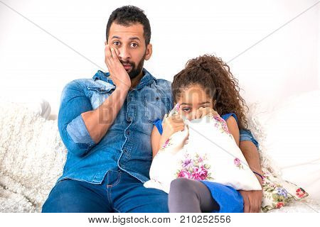 A young black father siting together with his daughter on the couch who is hiding behind a pillow and feeling scared