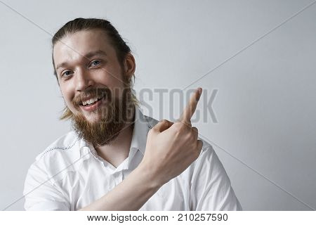 Headshot of positive friendly looking bearded businessman student or employee wearring earring and formal white shirt smiling broadly and indicating blank copyspace wall pointing index finger