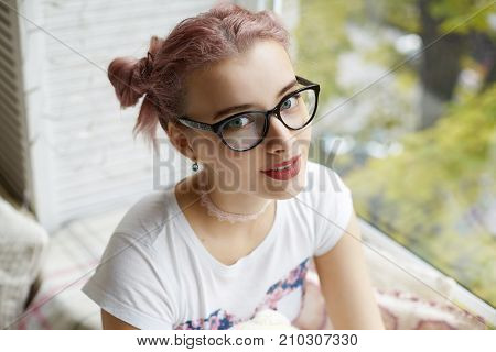 Close up portrait of beautiful trendy looking girl with pink colored hair sitting on plaid on windowsill relaxing after school staring at camera with her big blue eyes. Youth and happiness concept