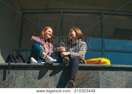 College students take a break after classes and chatting happy. Young girls with reddish hair sit on a modern building near windows on campus outdoors. Friendship and studentship.