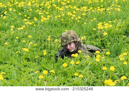 Portrait of armed woman with camouflage. Young female soldier observe with firearm. Child soldier with gun in war green goutweed and yellow dandelions background. Military army people concept