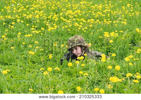 Portrait of armed woman with camouflage. Young female soldier observe with firearm. Child soldier with gun in war green goutweed and yellow dandelions background. Military army people concept