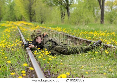 Portrait of armed woman with camouflage. Young female soldier observe with firearm on rail. Child soldier with gun in war green grass and yellow dandelions background. Military army people concept