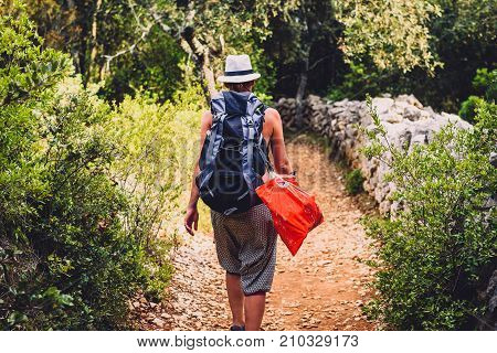 Woman hiking on country footpath with rock fence and woods. Hiker with a hat is trekking through rough jungle terrain on island Silba Croatia.