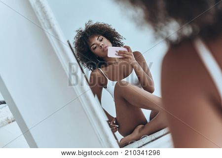 She will melt your heart. Reflection of beautiful young African woman in white lingerie taking selfie in the mirror while sitting on the floor at home
