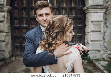 Happy Couple Of Newlyweds Posing Near Ancient Wooden Castle Gates, Handsome Smiling Groom Embracing