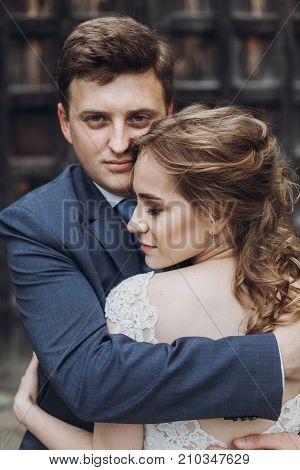 Happy Couple Of Newlyweds Posing Near Ancient Wooden Castle Gates, Handsome Smiling Groom Embracing