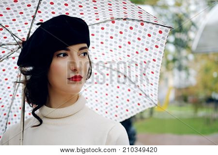 Young Parisian woman wearing black beret walking under the rain with dots umbrella. melancholic autumn mood.