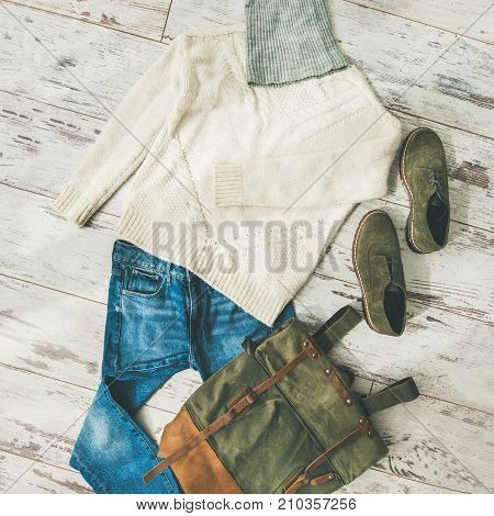 Flat-lay of Fall lady' s clothes outfit. Ecru colored knitted woolen sweater with braids, blue denim jeans, haki suede oxford boots, large backpack, cap over parquet background, top view, square crop
