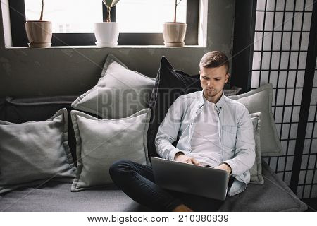 Handsome and attractive young man is sitting near bright window. He is keeping a laptop on his knees and working with it. This guy looks serious and thoughtful.