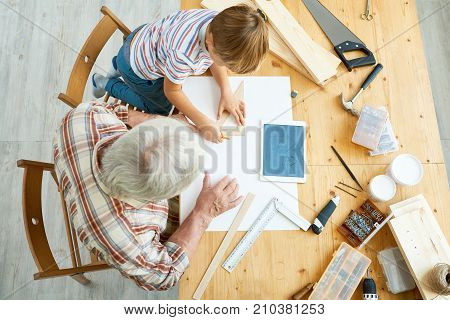 Above view portrait of little boy and granddad sitting together at desk sketching plan of birdhouse for school project and using digital tablet