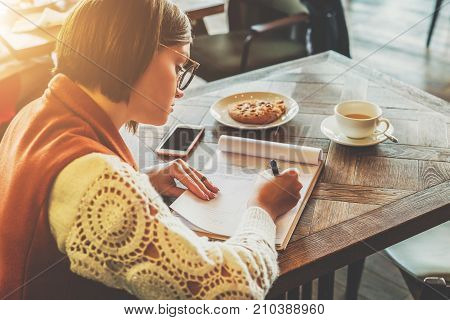 Side view. A young attractive woman sits in a cafe at a table and writes a pen, fills out a questionnaire, signs an application, compiles a CV. Online education, marketing. A student does homework.