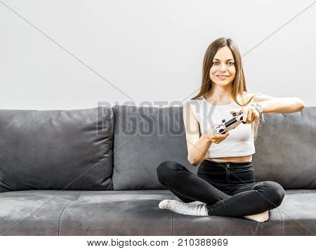 Pretty teenager girl playing a video game joyfully on the comfortable grey sofa before white wall