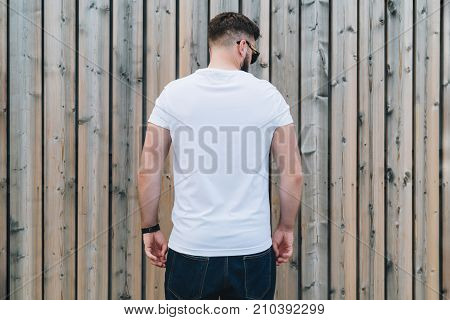 Summer day. Back view. Young bearded hipster man dressed in white t-shirt and sunglasses is stands outdoor against wood wall. Mock up. Space for logo, text, image.