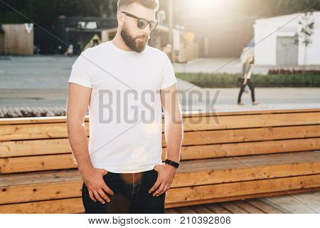Summer day. Front view. Young bearded hipster man dressed in white t-shirt and sunglasses is stands on city street. Mock up. Space for logo, text, image. Instagram filter, film effect, bokeh effect.