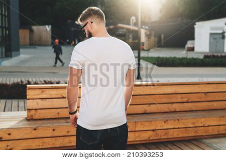 Summer day. Back view. Young bearded hipster man dressed in white t-shirt and sunglasses is stands on city street. Mock up. Space for logo, text, image. Instagram filter, film effect, bokeh effect.