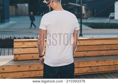 Summer day. Back view. Young bearded hipster man dressed in white t-shirt and sunglasses is stands on city street. Mock up. Space for logo, text, image. Instagram filter, film effect, bokeh effect.