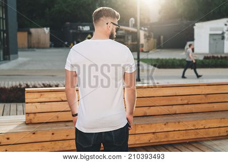 Summer day. Back view. Young bearded hipster man dressed in white t-shirt and sunglasses is stands on city street. Mock up. Space for logo, text, image. Instagram filter, film effect, bokeh effect.