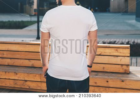 Summer day. Back view. Young bearded hipster man dressed in white t-shirt and sunglasses is stands on city street. Mock up. Space for logo, text, image. Instagram filter, film effect, bokeh effect.