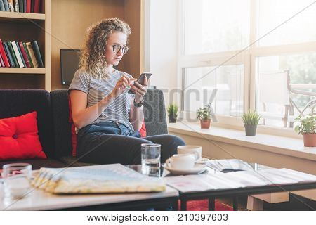 Young businesswoman sits on sofa near window, uses smartphone. Girl chatting, blogging, checking email, working. Student studying. Online marketing, education, e-learning, e-commercesocial medianetwork