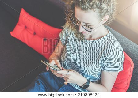View from above. Young woman in glasses and a striped T-shirt is sitting at home on the couch and is using a smartphone. Hipster girl is checking e-mail, chatting, blogging, messaging. Social network.