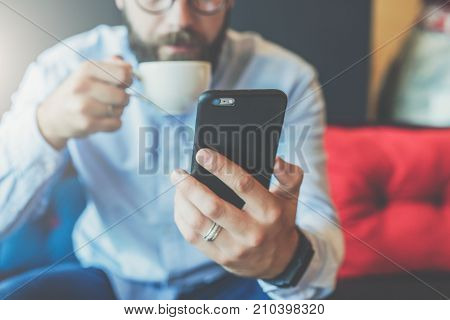 Close-up of black smartphone in man's hand. In background, in soft focus, young bearded businessman drinks coffee. Guy uses digital gadget, checking email, reading, blogging. Online working, education.