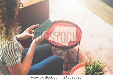 View from above. Young woman in glasses and striped T-shirt is sitting at home on couch and is using smartphone. Hipster girl is checking e-mail, chatting, messaging. Blurred background, film effect.
