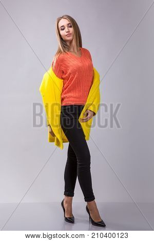 Elegance blonde girl is posing in the studio on the gray background. She wears black pants, orange pulover and yellow coat.