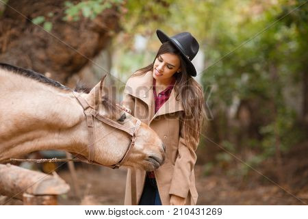 An attractive lady in cowboy hat with light brown horse on a blurred background. A young woman and a horse. People and animals concept.