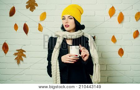 Woman with autumn leaves in scarf and hat. Fashion model with red lips makeup at brick wall. Girl with milk or hot wine in tea cup. Beauty and fall fashion. Season and autumn holiday.