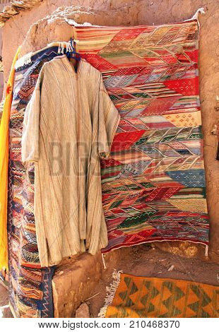 Traditional men's Moroccan clothing djellaba and wool carpet on clay wall, souk in Ait-Ben-Haddou Kasbah, Morocco, North Africa