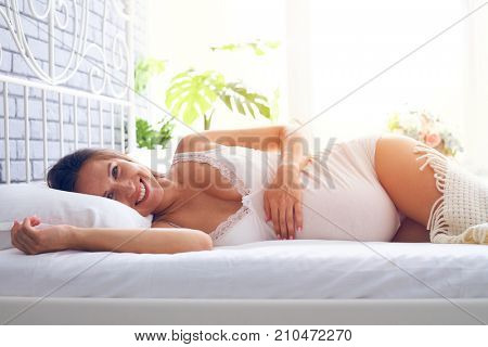 Closeup of smiling pregnant woman lying on side on the bed, holding hand on pregnant belly 