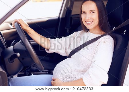 Side view of happy future mother with hand on belly posing on driver seat 