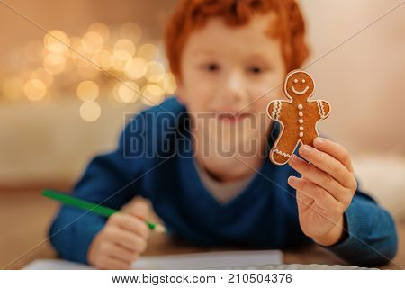 Look what I have. Selective focus on a tasty homemade gingerbread man held buy a smiling kid lying on the floor and drawing with a smile on his face.