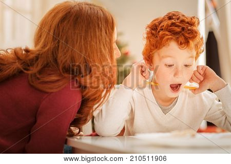 Cannot believe my eyes. Adorable curly haired boy keeping his mouth wide opened while getting surprised during a conversation with his mother.